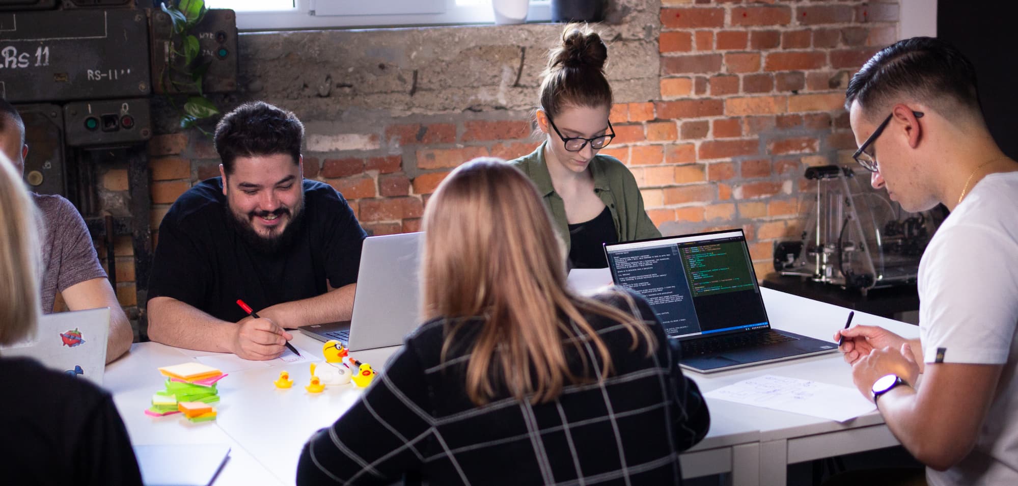 A project planning meeting at Revolve Healthcare office A group of people sitting at a long table with laptops and some rubber ducks on it, writing on A4 sheets of paper during a project planning meeting at an office with red brick wall